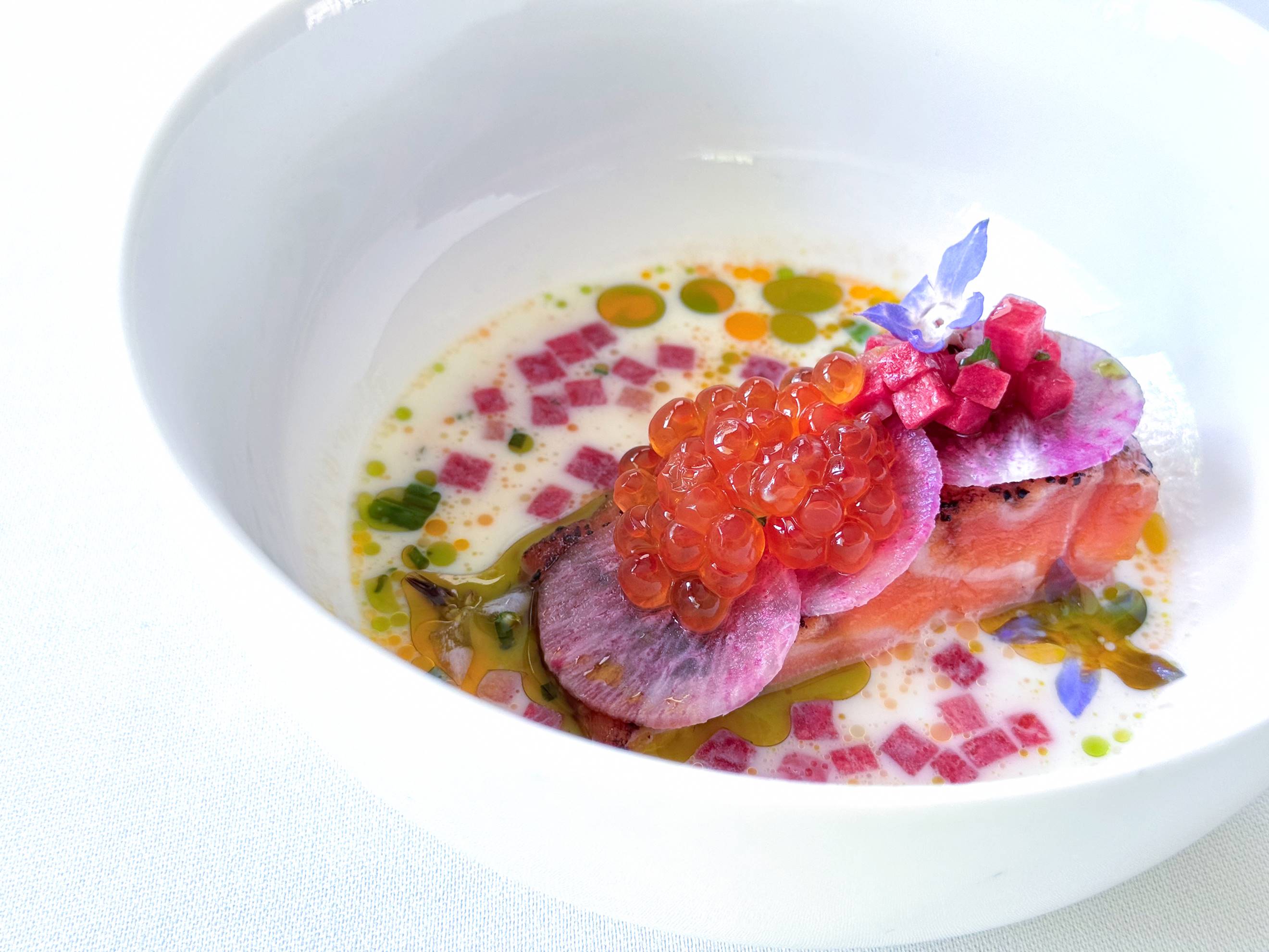 A white bowl with a gourmet dish featuring a slice of cooked salmon, topped with salmon roe, thinly sliced radish, pink diced garnish, edible flowers, and colorful oil drops in a creamy white sauce.