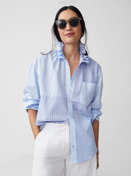 A woman wearing black sunglasses, a light blue and white striped button-up shirt with ruffled collar, and white pants stands against a plain light background, smiling slightly with her hands in her pockets.
