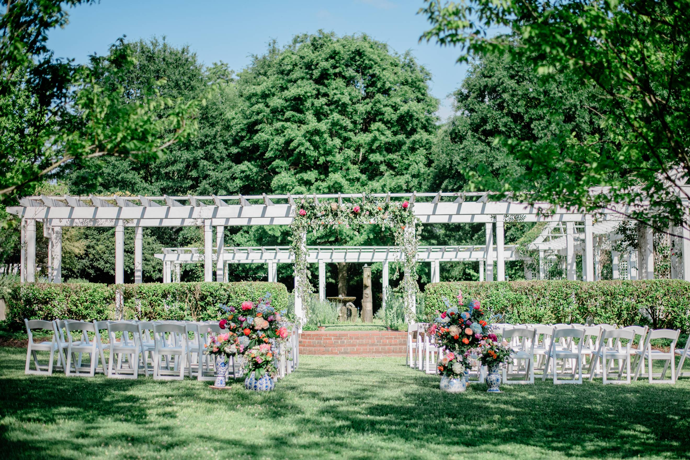 An outdoor wedding ceremony setup with rows of white chairs facing a pergola decorated with greenery and colorful flowers, surrounded by lush trees and greenery on a sunny day.