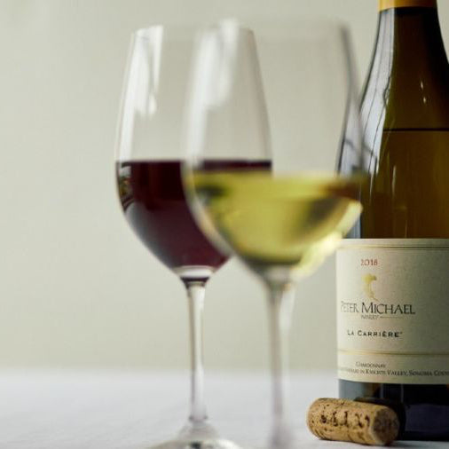 Two wine glasses, one filled with red wine and the other with white wine, stand beside a bottle of Peter Michael white wine and a cork on a white surface, with a light background.