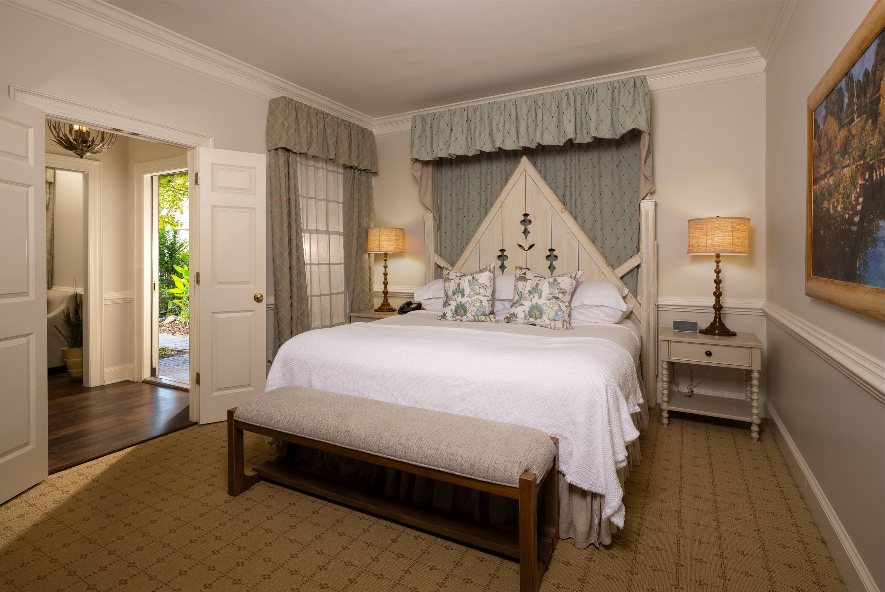 A cozy bedroom with a large bed featuring decorative pillows, two bedside tables with lamps, a cushioned bench at the foot of the bed, patterned curtains, and a door open to a hallway leading outside.