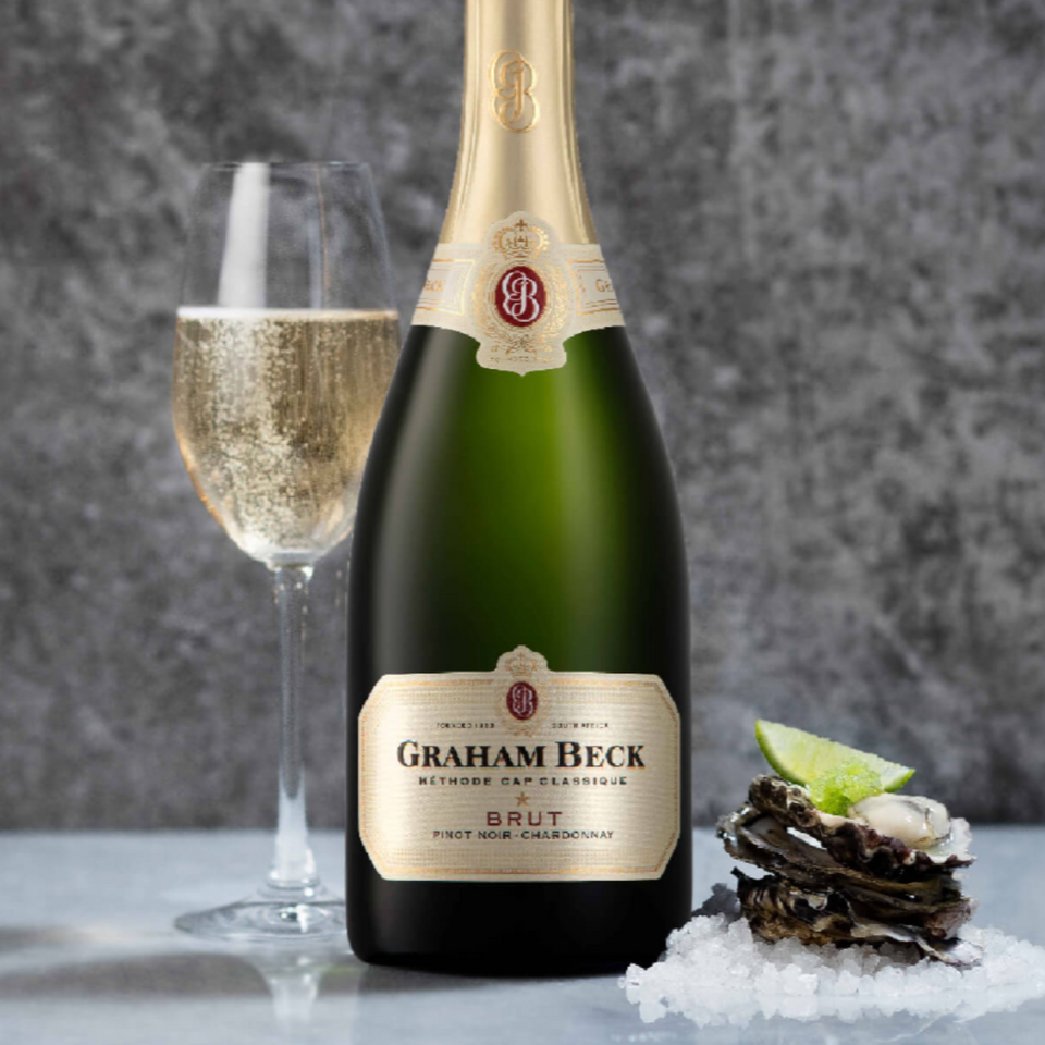 A bottle of Graham Beck Brut sparkling wine stands beside a filled champagne flute, with a stack of oysters on ice garnished with a lime wedge, set against a gray marbled background.