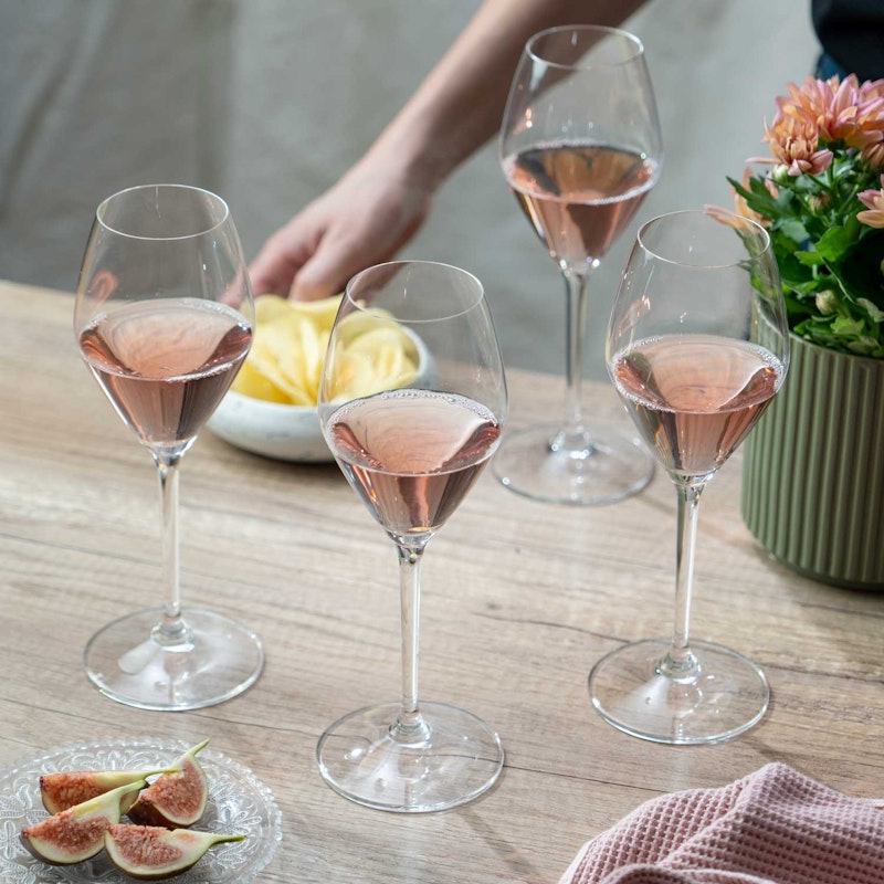 Four glasses of rosé wine on a wooden table, next to a plate of sliced figs, a pink napkin, a potted plant with flowers, and a hand reaching for a bowl of potato chips in the background.