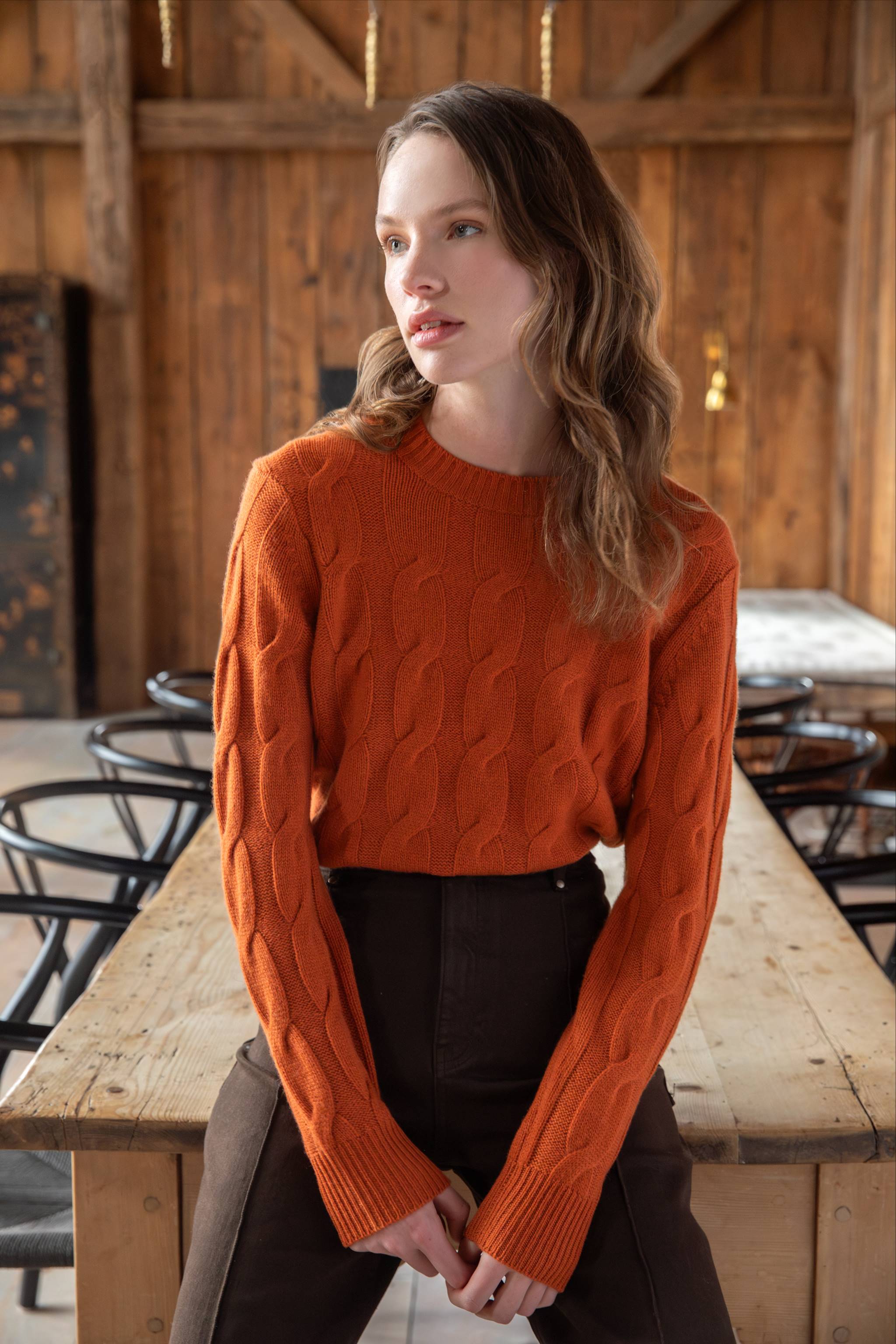 A woman with wavy light brown hair wearing an orange cable-knit sweater and dark pants sits on the edge of a wooden table in a rustic room with wooden walls and black chairs.