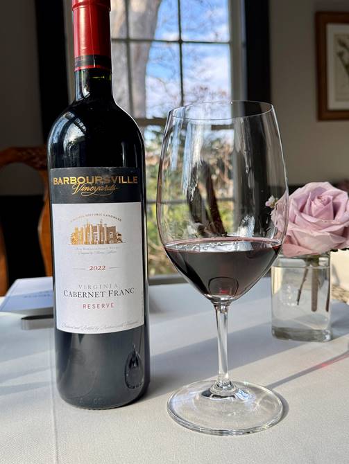A bottle of Barboursville Vineyards 2022 Virginia Cabernet Franc Reserve stands next to a half-filled wine glass on a white table. A vase with light pink roses and a window with a tree view appear in the background.