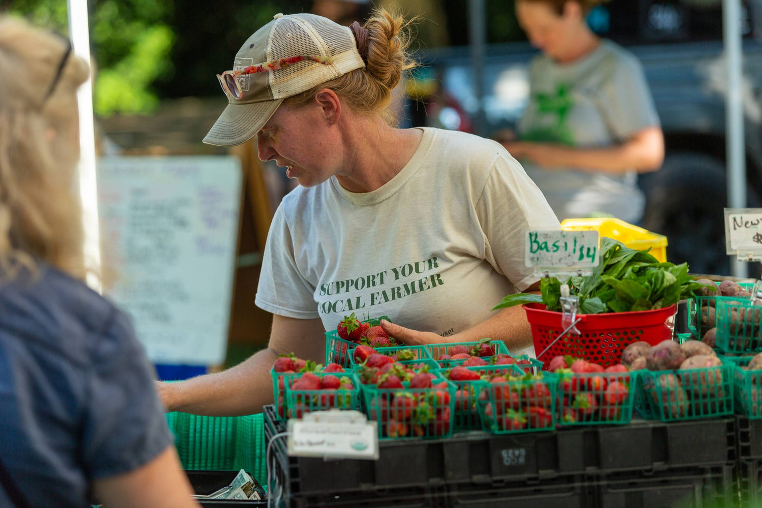 A woman wearing a cap and a "Support Your Local Farmer" t-shirt sells strawberries and produce at an outdoor market stand, arranging baskets of fresh berries and greens. Signs label the items as basil and new potatoes.