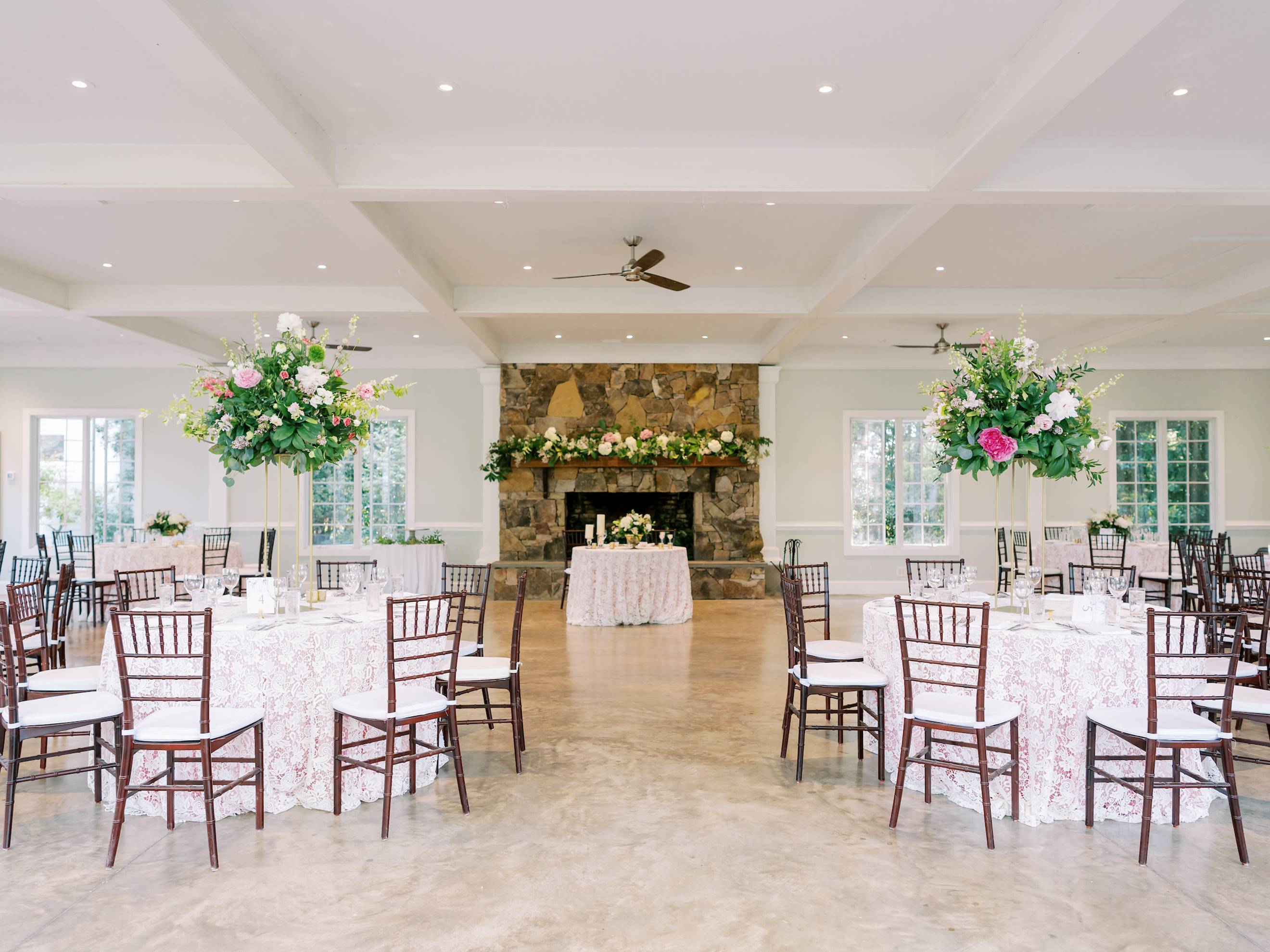A bright, elegant banquet hall with round tables covered in floral tablecloths, tall flower centerpieces, dark wooden chairs, and a large stone fireplace decorated with greenery and flowers.