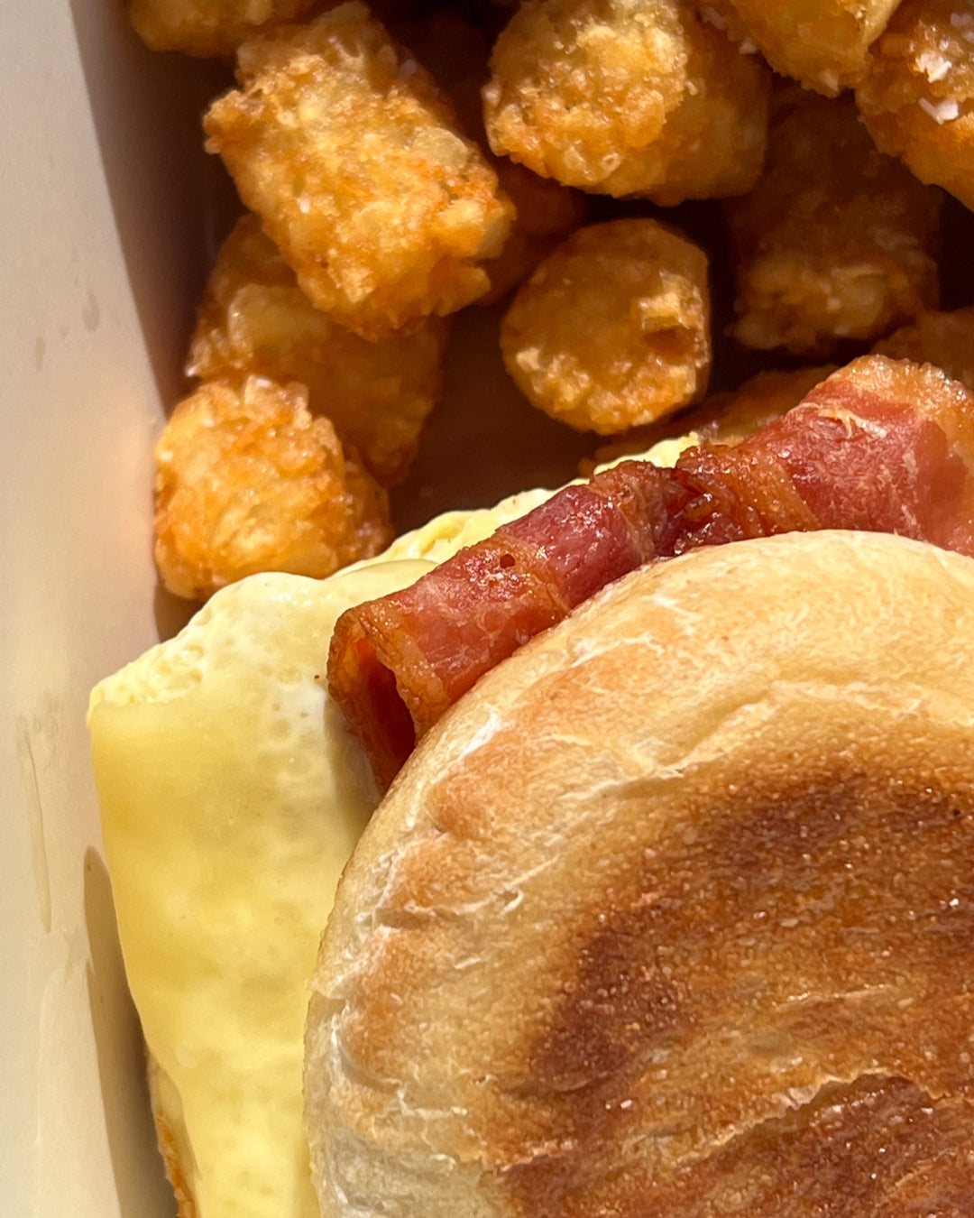 A close-up of a breakfast sandwich with bacon, melted cheese, and an English muffin, next to a pile of crispy tater tots in a takeout container.