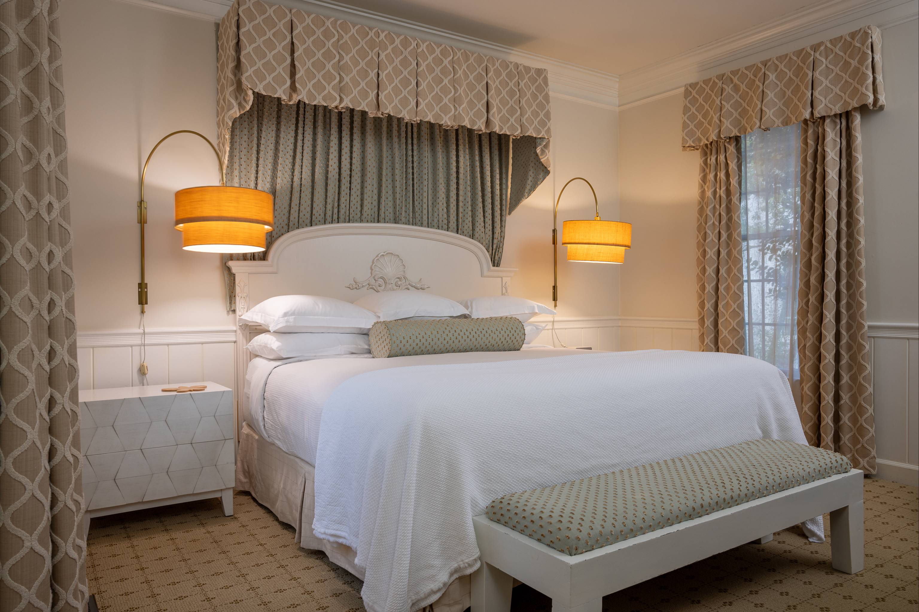A cozy bedroom with a white bed, patterned pillows and bench, matching curtains, two wall-mounted lamps with yellow shades, and light-colored walls and carpet. Sunlight streams through a window on the right.