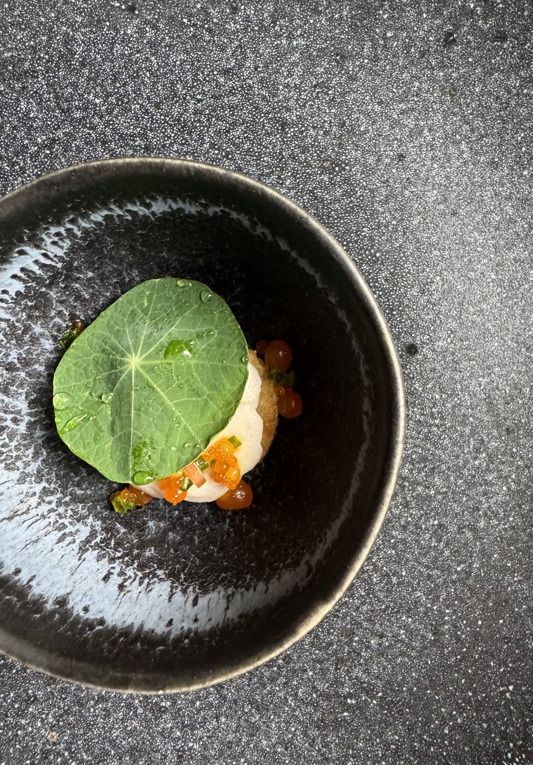 A black bowl with a gourmet dish featuring a green leaf topping a mound of food, garnished with orange roe and herbs, set against a textured dark surface.