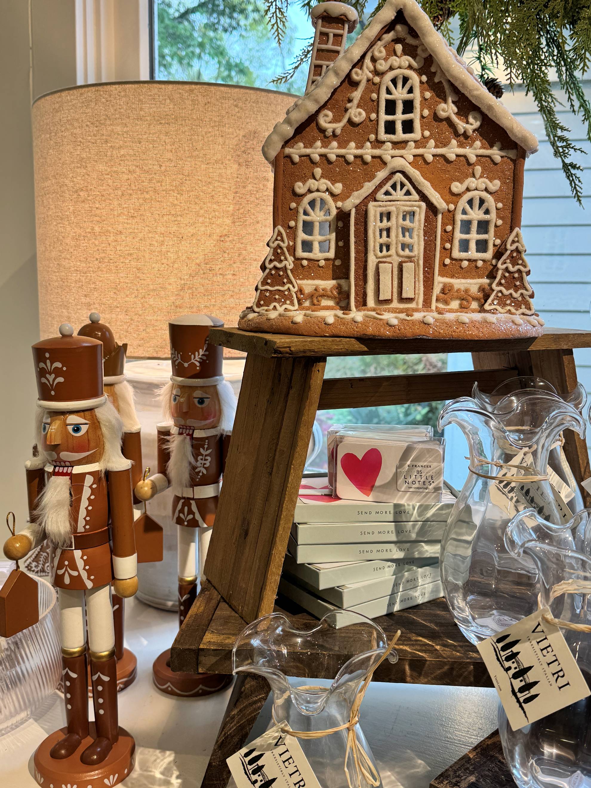 A tabletop display features brown nutcracker figurines, a decorative gingerbread house, glass pitchers, stacked gift boxes, and a lit lamp, all set near a window with greenery overhead.
