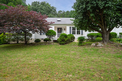 Single-story white house with a gray roof and black shutters, surrounded by green bushes and trees, with a grassy lawn in the foreground and a small covered porch by the front door.