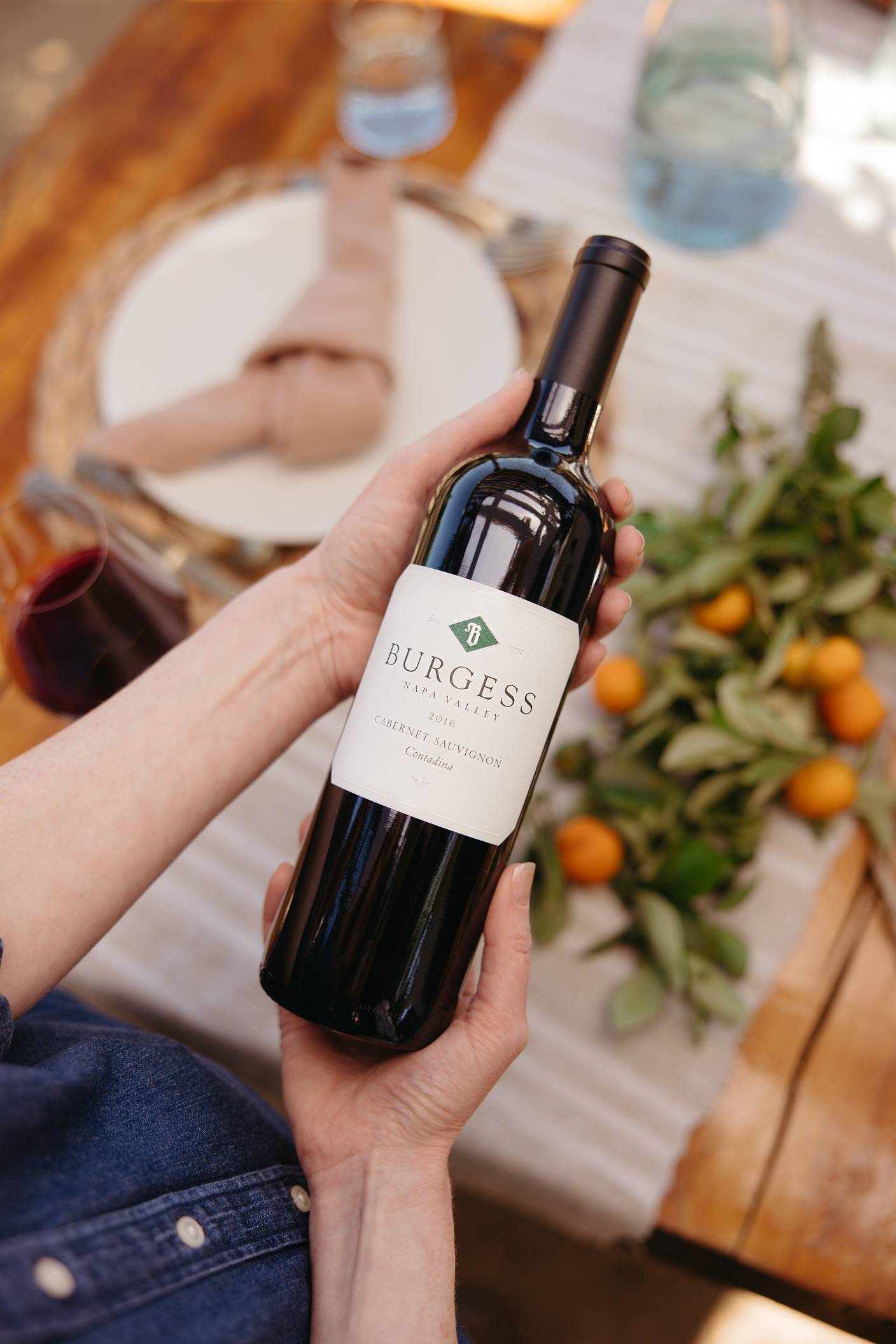 A person holds a bottle of Burgess Cabernet Sauvignon wine over a table set with a plate, napkin, water glass, and a centerpiece of leafy branches with small orange fruits.
