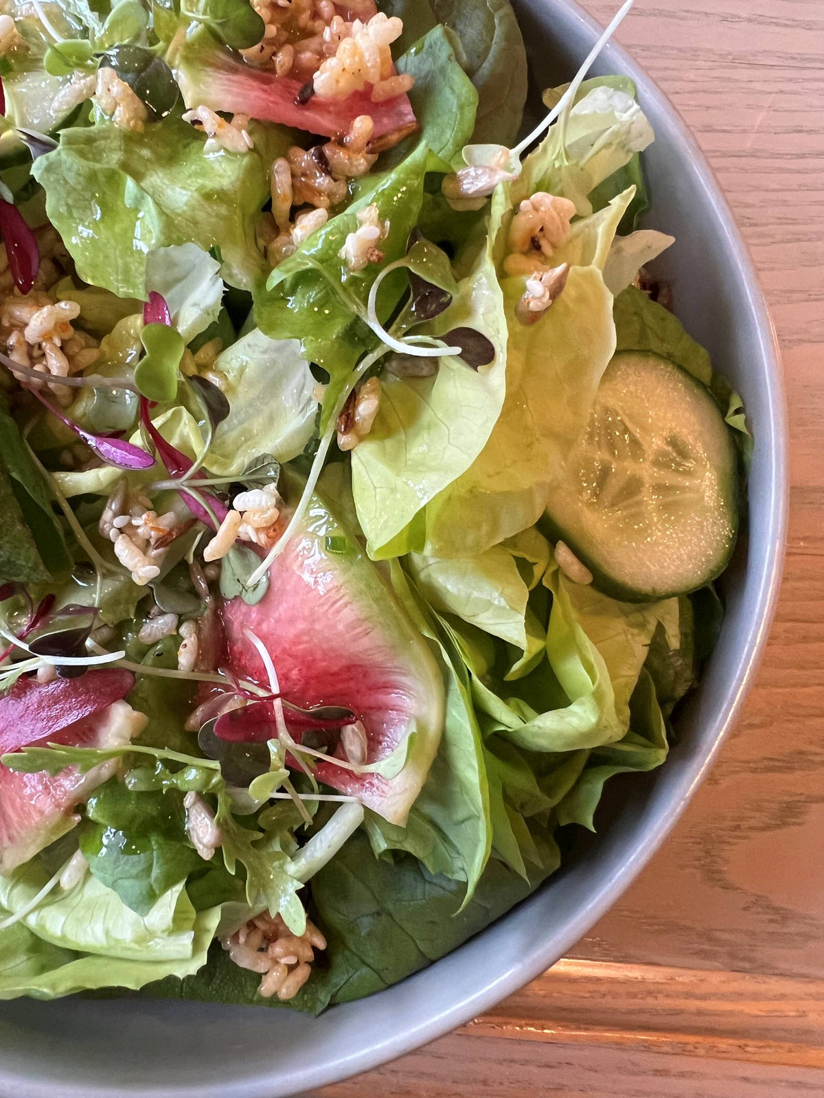 A close-up of a fresh salad in a bowl, featuring mixed greens, cucumber slices, microgreens, rice crisps, and sliced figs on a wooden table.