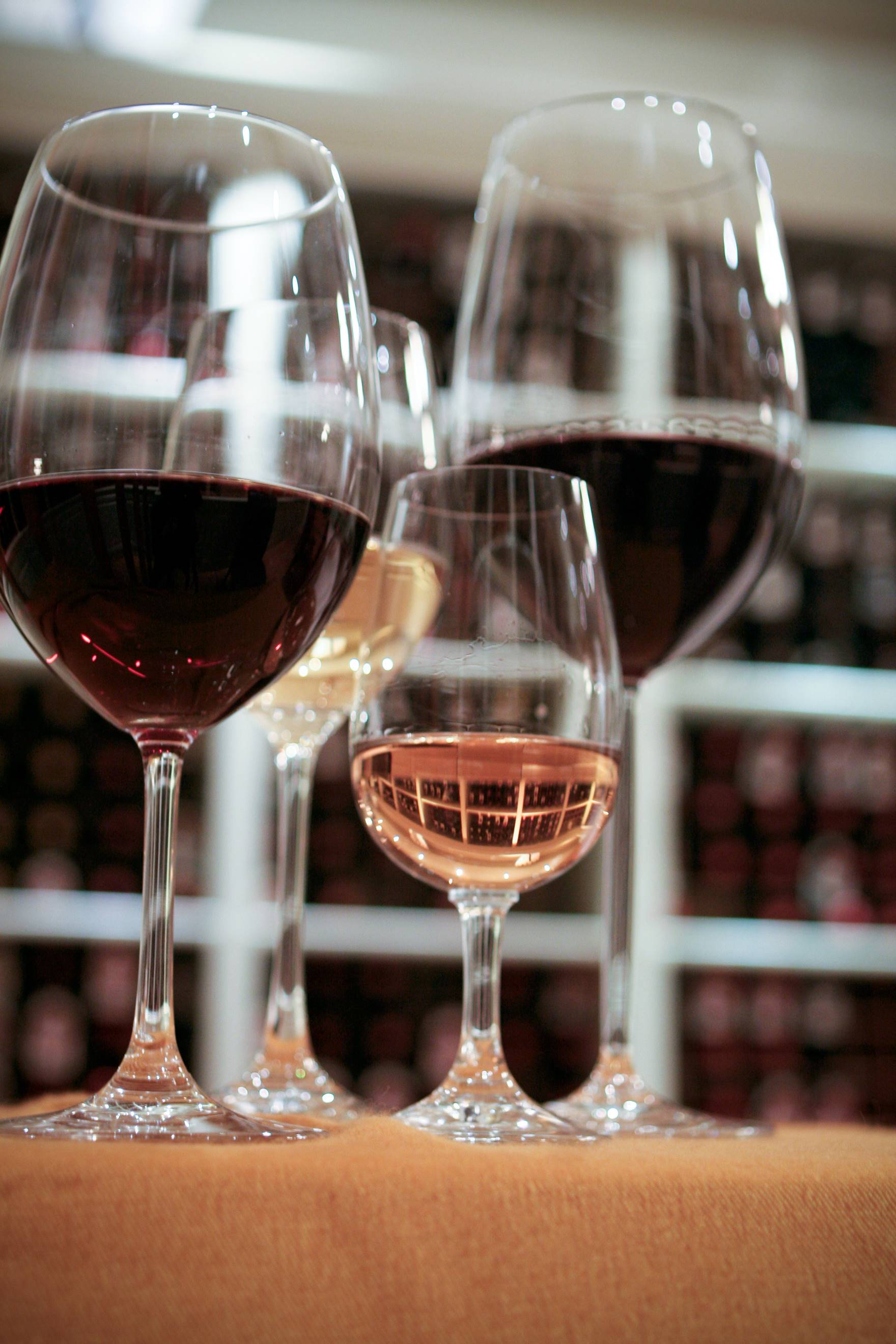 Four wine glasses containing red, white, and rosé wines are lined up on a brown surface, with wine racks blurred in the background.
