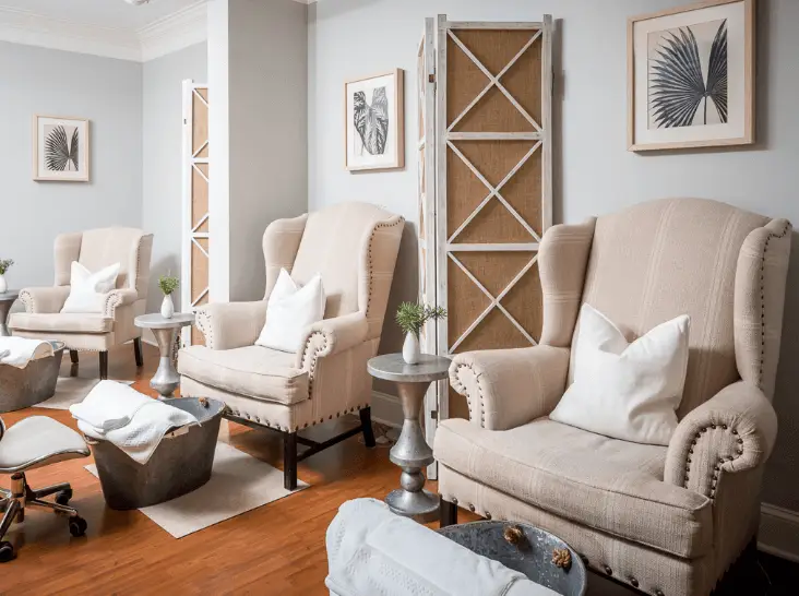 A spa lounge with three beige armchairs, white pillows, silver side tables, foot basins, and framed leaf artwork on light gray walls. Wood floors and a decorative room divider add to the calming, elegant atmosphere.