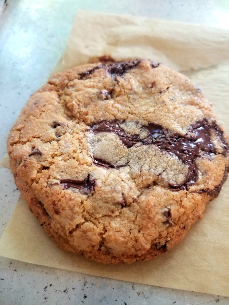 A close-up of a freshly baked chocolate chip cookie resting on a piece of parchment paper, with melted chocolate visible on the cookie's surface.