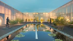 A modern courtyard with a reflective rectangular pond filled with water lilies, surrounded by sculptures, plants, gravel paths, and buildings with translucent walls softly illuminated at dusk.
