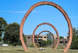 Three large, rust-colored metal rings stand upright in a grassy field, aligned in a row. A modern building with tan walls and glass windows is visible in the background under a clear blue sky.