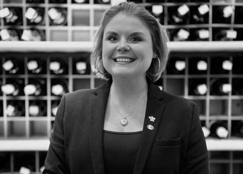 A woman in a blazer smiles at the camera, standing in front of shelves filled with wine bottles. She wears a necklace and pins on her lapel. The photo is in black and white.