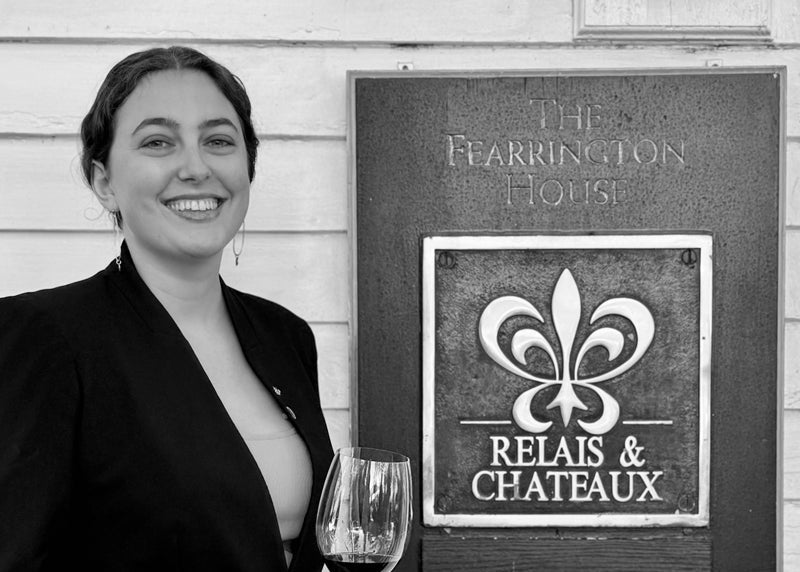 A smiling woman in a dark blazer holds a wine glass and stands next to a sign reading "The Fearrington House" and "Relais & Chateaux" with a fleur-de-lis logo. The image is in black and white.