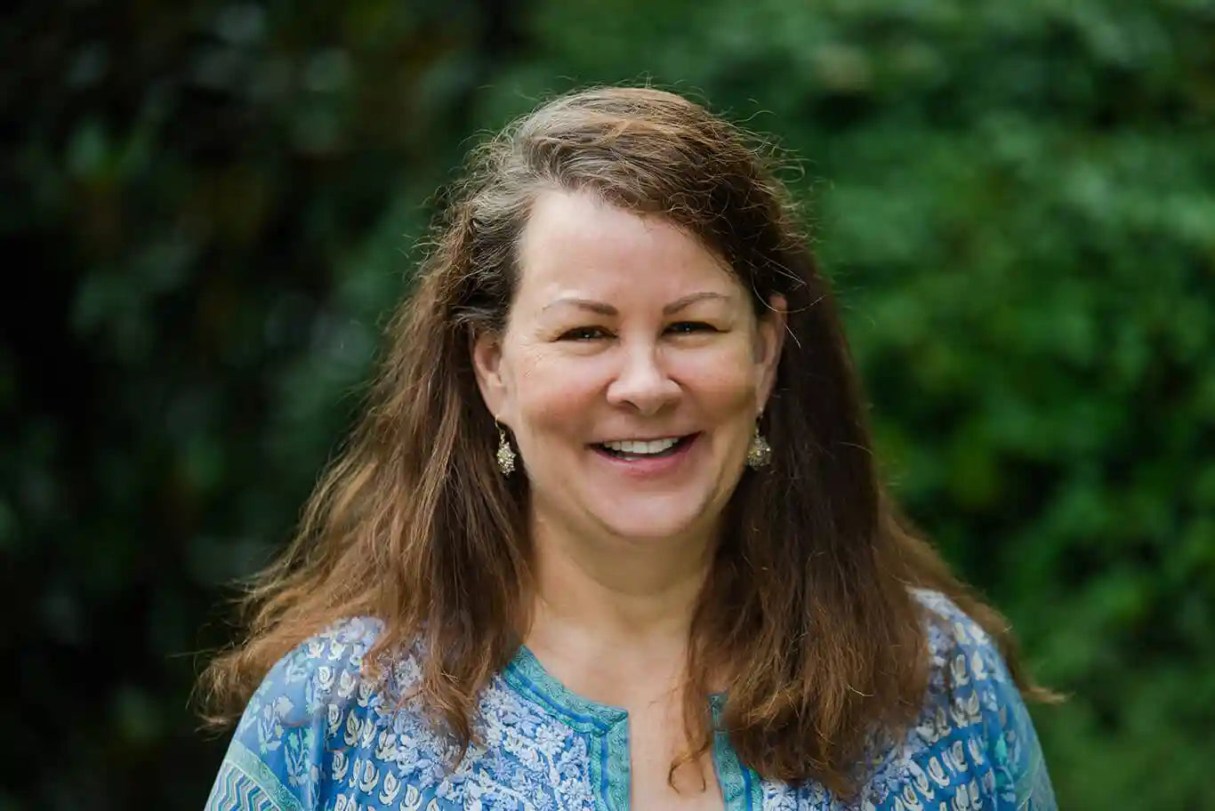 A woman with long brown hair wearing a blue patterned blouse and dangling earrings smiles outdoors in front of a blurred green background.