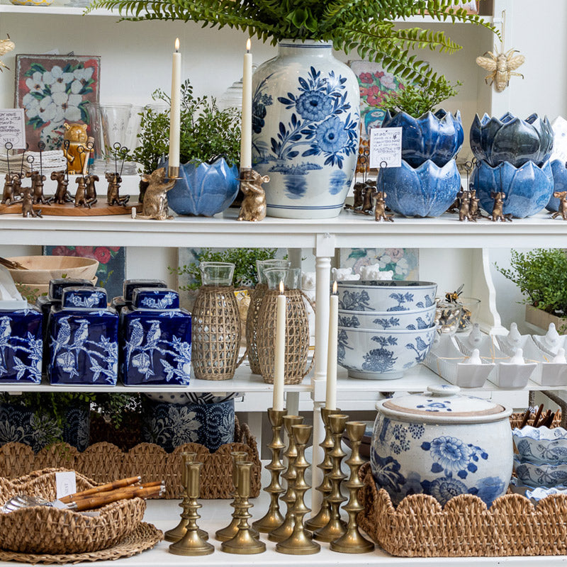 A display of blue and white ceramic vases, bowls, and candle holders on white shelves, accented with brass candlesticks, wicker trays, small plants, and decorative figurines in a home decor shop.