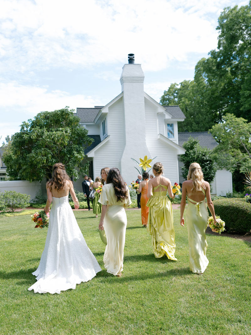 Five women in pastel dresses, including a bride in white, walk across a grassy yard toward a white house, surrounded by greenery and flowers under a blue sky.