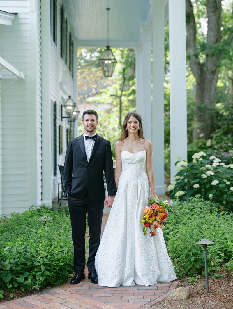 A bride in a white gown holding a colorful bouquet stands beside a groom in a black tuxedo on a brick path, surrounded by greenery near a white building.