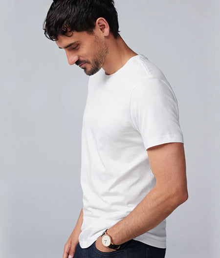 A man with short dark hair wearing a plain white t-shirt and jeans stands in profile against a light gray background, looking down with a slight smile. He has a watch on his left wrist.