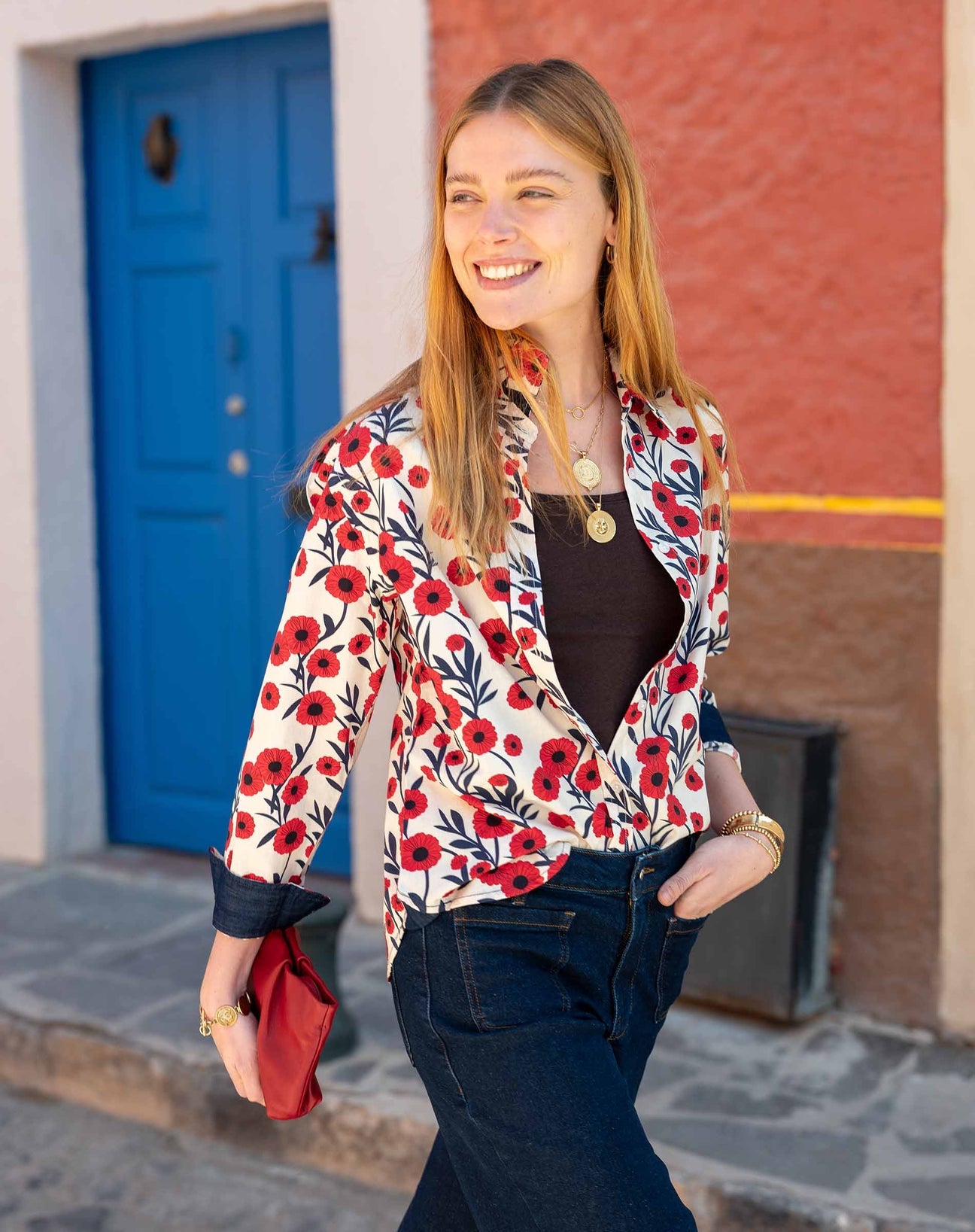 A smiling woman with long blonde hair walks outside, wearing a white shirt with a red floral pattern, dark jeans, gold jewelry, and holding a small red bag. A blue door and colorful walls are in the background.