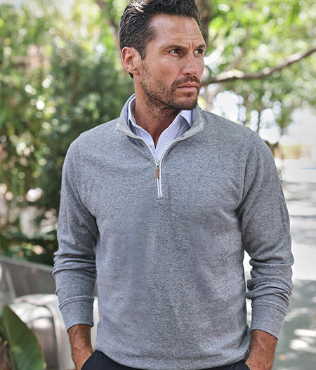 A man wearing a light gray quarter-zip sweater over a collared shirt stands outdoors, looking to the side. Trees and greenery are in the blurred background, with sunlight filtering through the leaves.