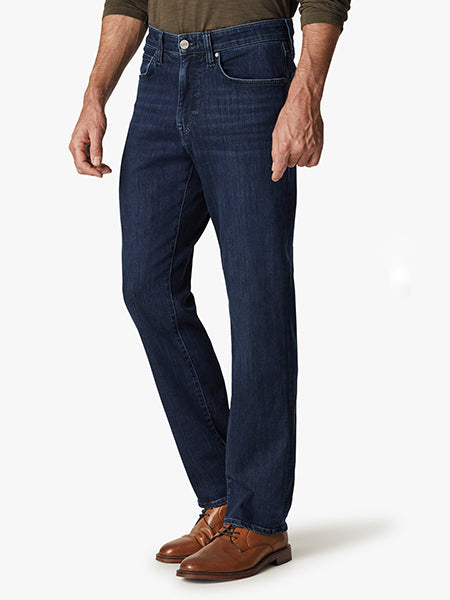 A person wearing dark blue straight-leg jeans, a long-sleeve olive green shirt, and brown leather dress shoes stands against a plain white background.