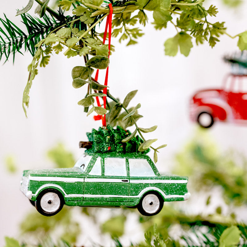 A green car ornament with a tiny Christmas tree on its roof hangs from a Christmas tree branch, with blurred greenery and a red car ornament in the background.