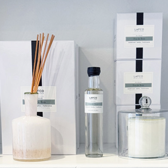 A display of LAFCO home fragrance products, including a reed diffuser in a white ceramic bottle, a clear bottle of fragrance oil, boxed items, and a white candle with a glass lid, arranged on a white shelf.
