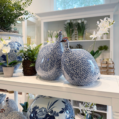 Two blue and white ceramic guinea fowl figurines with dotted patterns sit on a white shelf, surrounded by potted plants, orchids, and decorative vases in a bright, elegant interior.