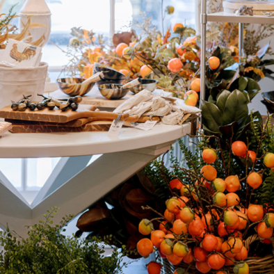 A round table displays wrapped utensils, bowls, and wooden boards, surrounded by lush greenery and vibrant orange artificial fruit arrangements in a bright, inviting setting.
