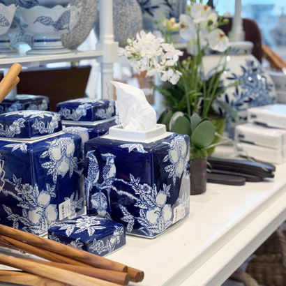 A display of blue and white ceramic jars with floral patterns, one holding tissues, surrounded by wooden utensils, artificial white orchids, and other decorative items on a white table.