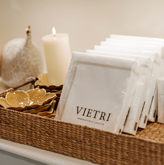 A wicker tray holds yellow ceramic bowls, a white pillar candle, and several packages of VIETRI branded paper napkins. A decorative speckled white object is placed behind the candle.