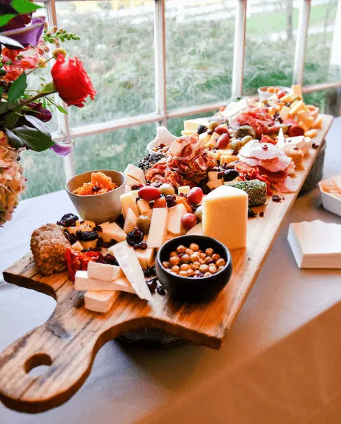A wooden board on a table holds an assortment of cheeses, cured meats, nuts, berries, and crackers near a bright window, with flowers and napkins nearby.