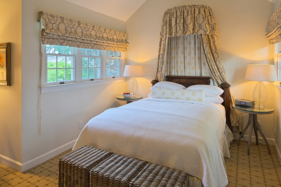 A cozy bedroom with a double bed covered in white bedding, two wicker benches at the foot, matching side tables with lamps, patterned curtains, and windows letting in natural light.