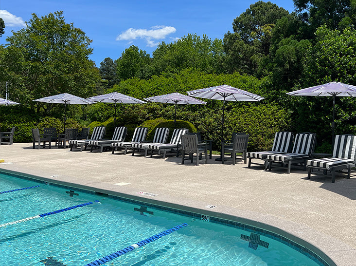 A row of black-and-white striped lounge chairs and umbrellas lines the side of an outdoor swimming pool, surrounded by greenery and trees under a sunny blue sky.