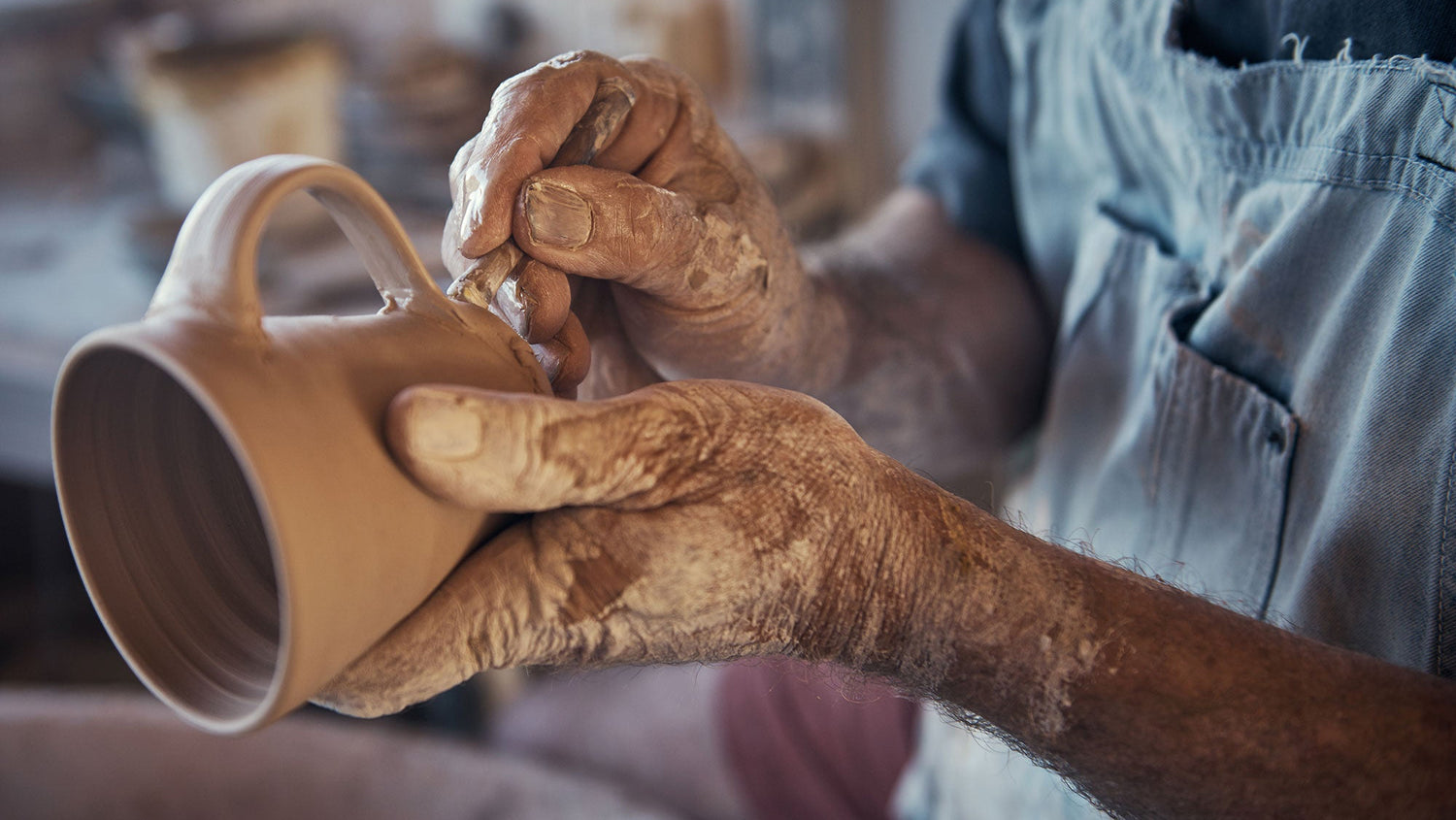 A person with clay-covered hands shapes the handle of a ceramic mug, wearing a blue apron and working in a pottery studio.