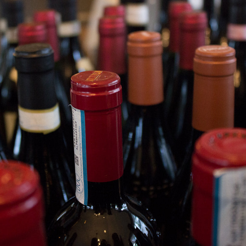 Close-up of the tops of multiple wine bottles arranged closely together, featuring various colored caps including red, black, and brown. The focus is on a red-capped bottle in the center.
