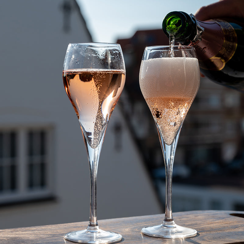 Two elegant champagne glasses sit on a table; one is filled with sparkling rosé, while the other is being filled with bubbly champagne from a green bottle, with bubbles rising in both glasses.
