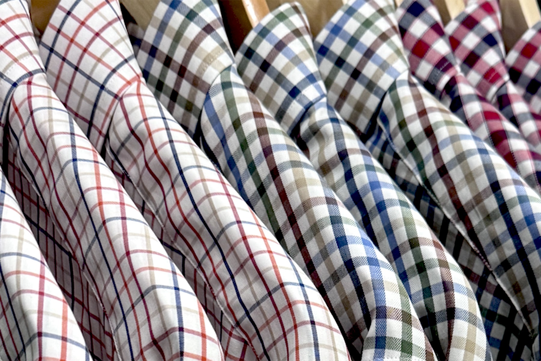A row of collared shirts with various plaid patterns and colors, neatly hung on wooden hangers. The shirts feature different combinations of lines in red, blue, green, and brown on white backgrounds.