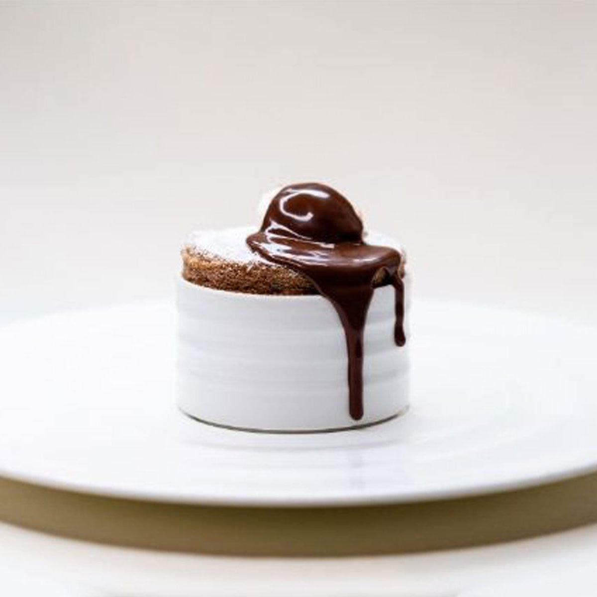 A chocolate soufflé in a white ramekin is topped with a scoop of ice cream and rich chocolate sauce, which drips down the side, set on a white plate against a plain background.