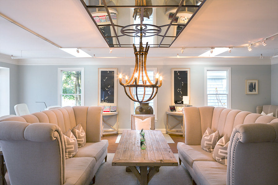 A modern living room with high-back beige sofas around a wooden table, a large metal chandelier, wall art, and a mirrored ceiling panel reflecting the light fixture. Natural light enters through windows.