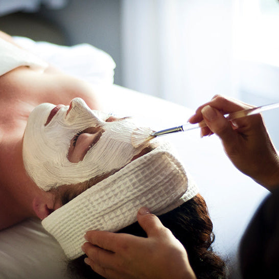 A person lies on a spa table with eyes closed while an esthetician applies a white facial mask using a brush. The person is wearing a white towel headband.