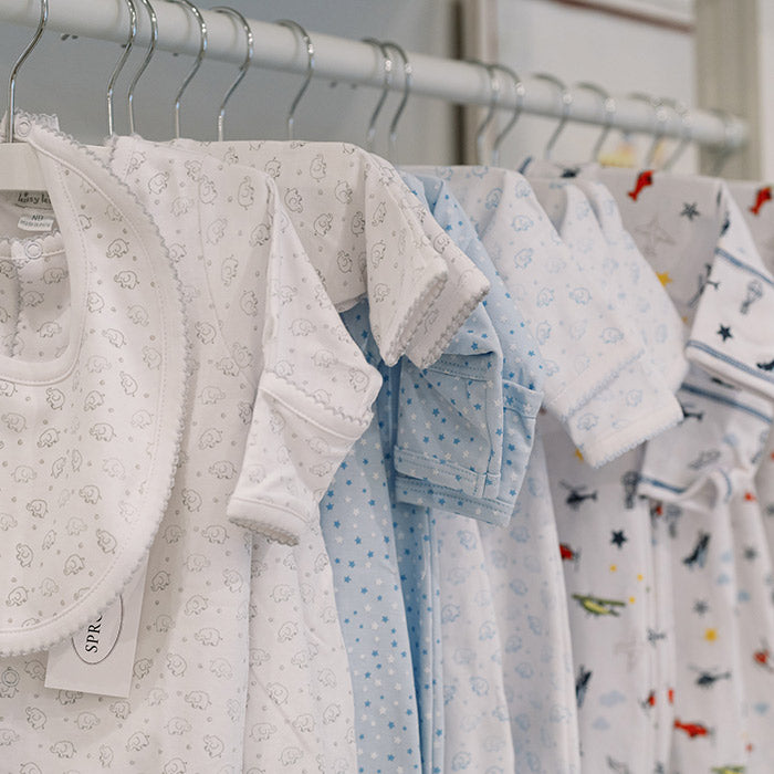 A row of baby clothes and bibs in soft pastel colors, mainly white and blue, with small patterns like animals and stars, hanging on hangers on a clothing rack.